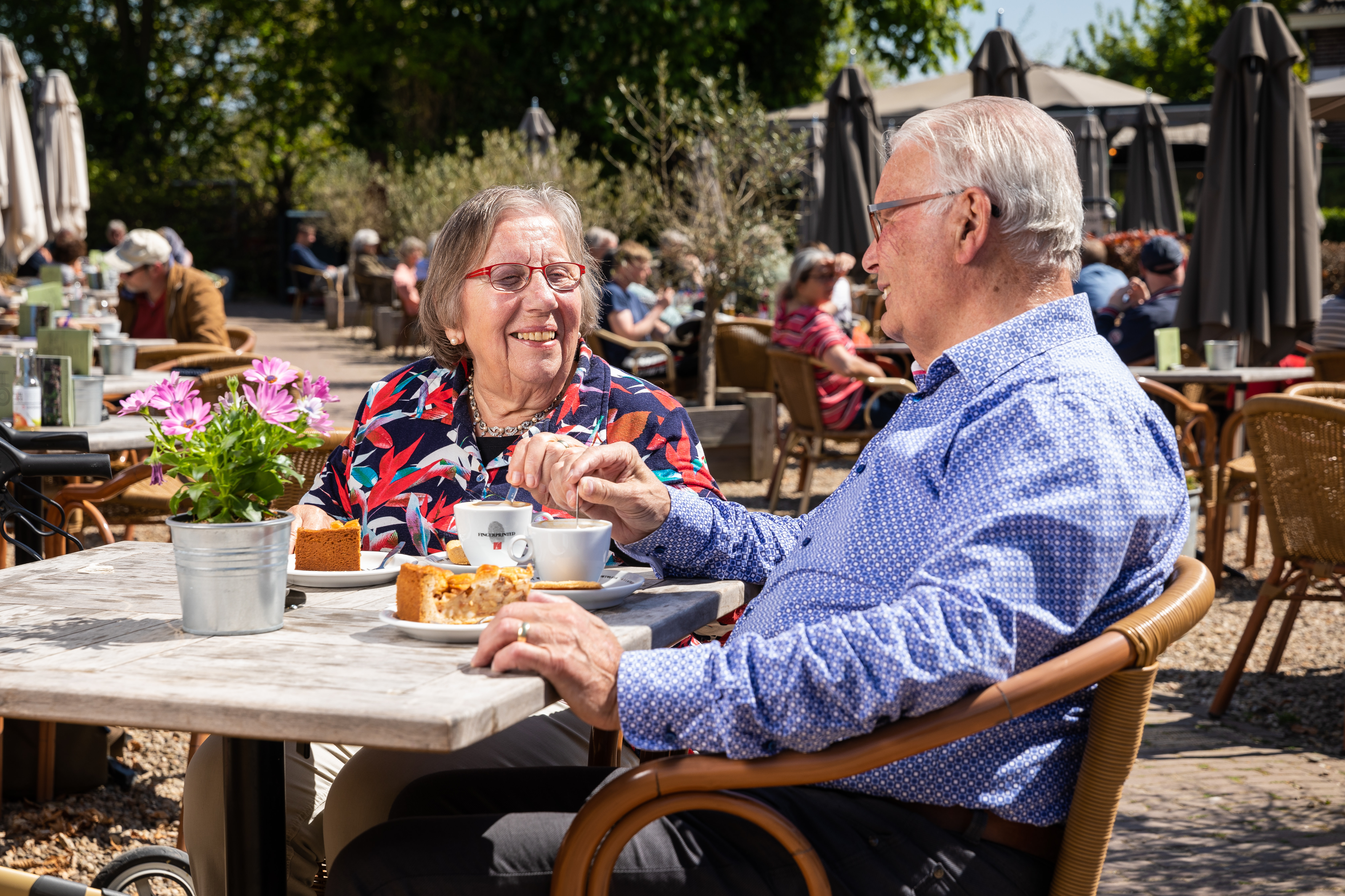Ouder echtpaar zit op een zonnig terras met koffie en appeltaart.