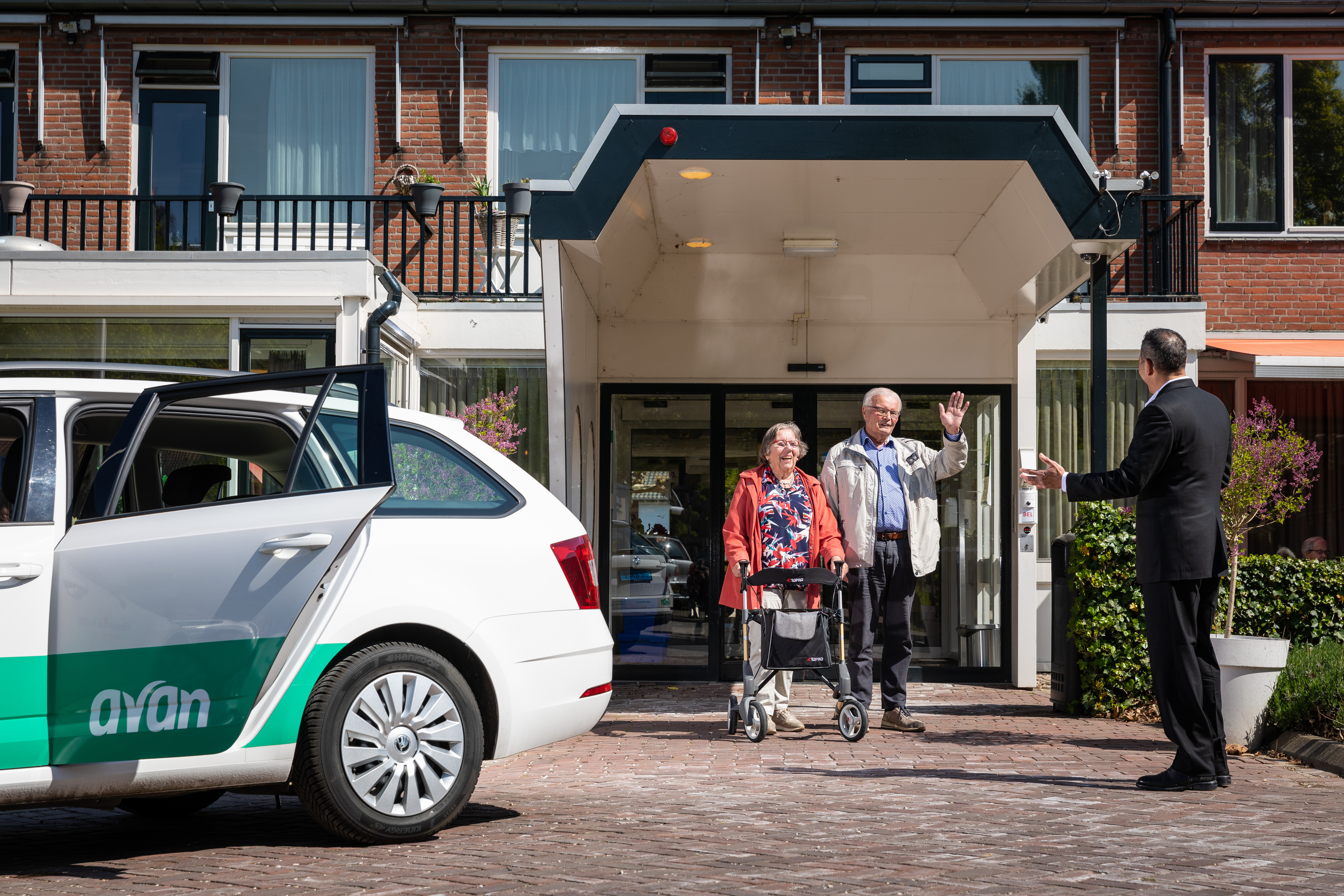 Echtpaar loopt naar buiten bij de woning en zwaait naar de Avan chauffeur hen op komt halen met een personenauto.