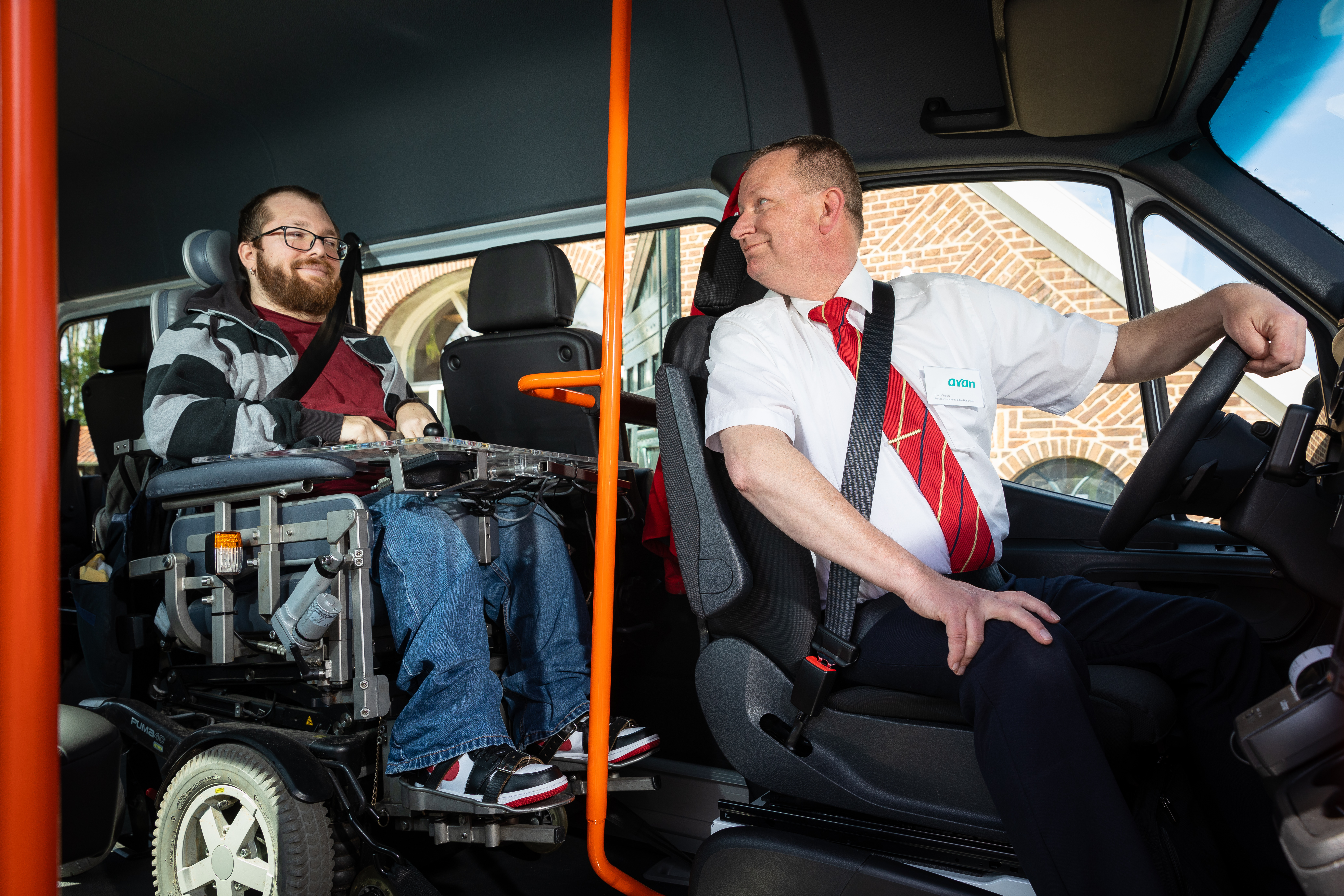 Een reiziger in een rolstoel zit in de taxibus, de chauffeur kijkt om naar de reiziger.