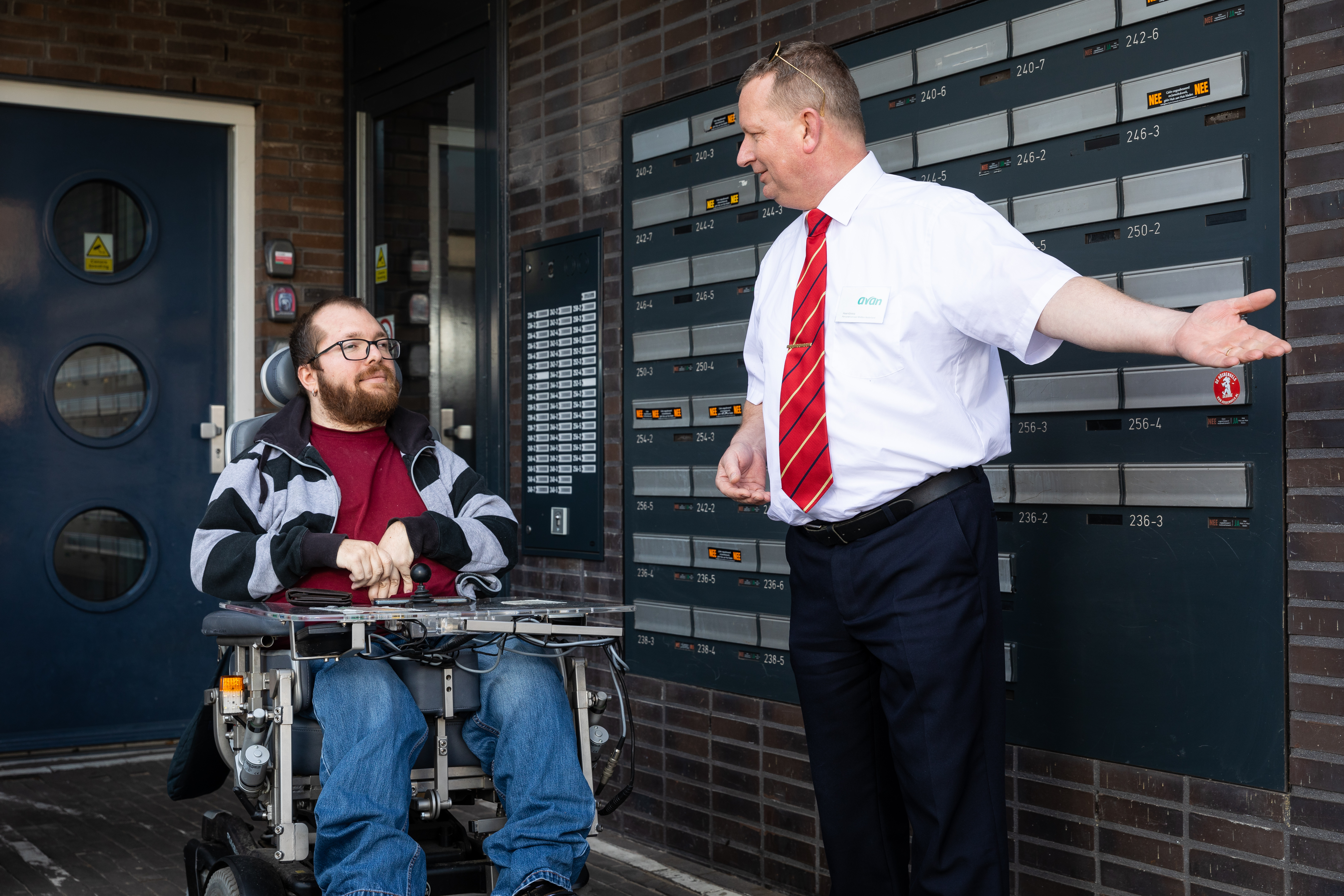 Een chauffeur heet een een reiziger in rolstoel welkom beneden bij een flatgebouw.